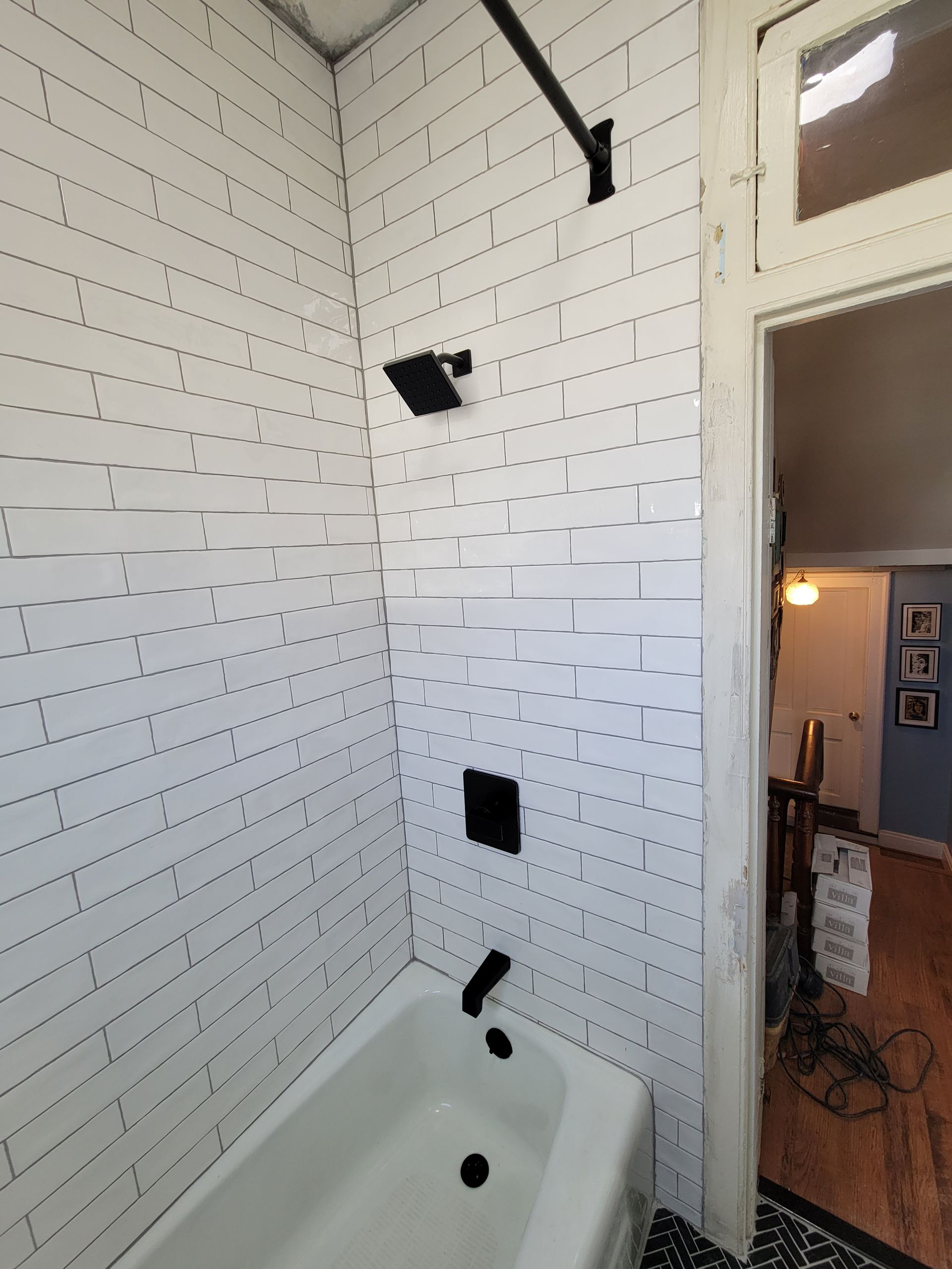 White tiled shower corner with black fixtures and a tub, viewed through an open bathroom door