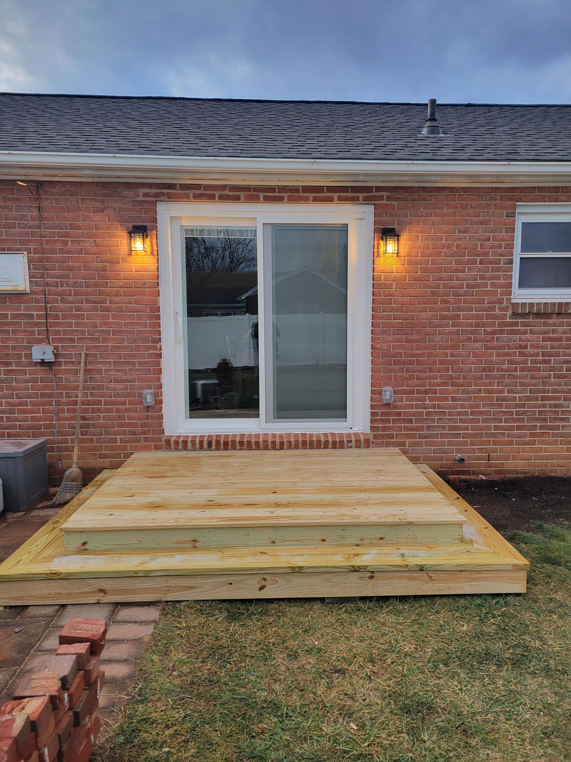 Backyard deck under construction behind a brick house with sliding glass doors and lit wall sconces
