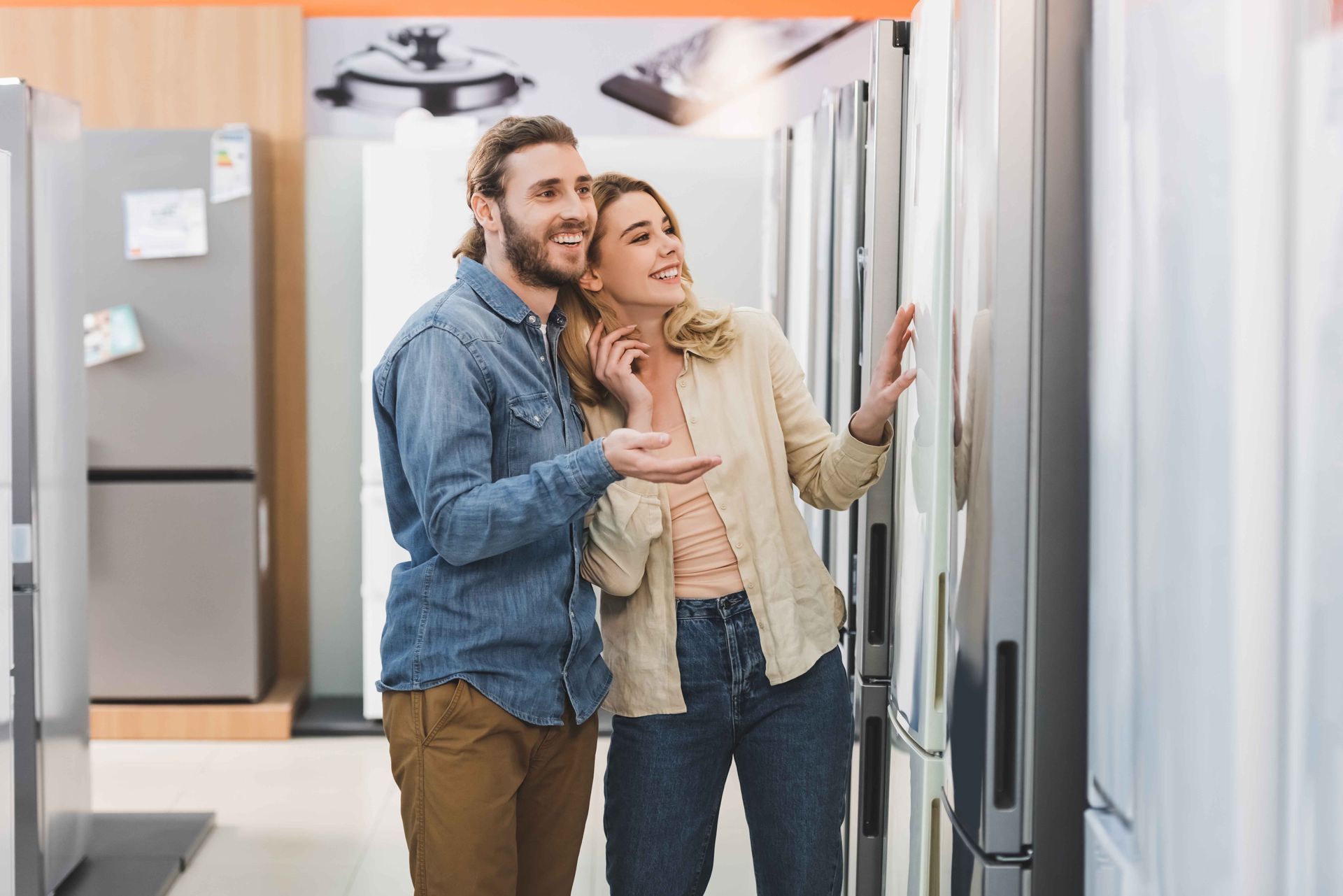 Un couple fait ses achats de réfrigérateurs dans un magasin d'électroménager, souriant et montrant du doigt les différents modèles.
