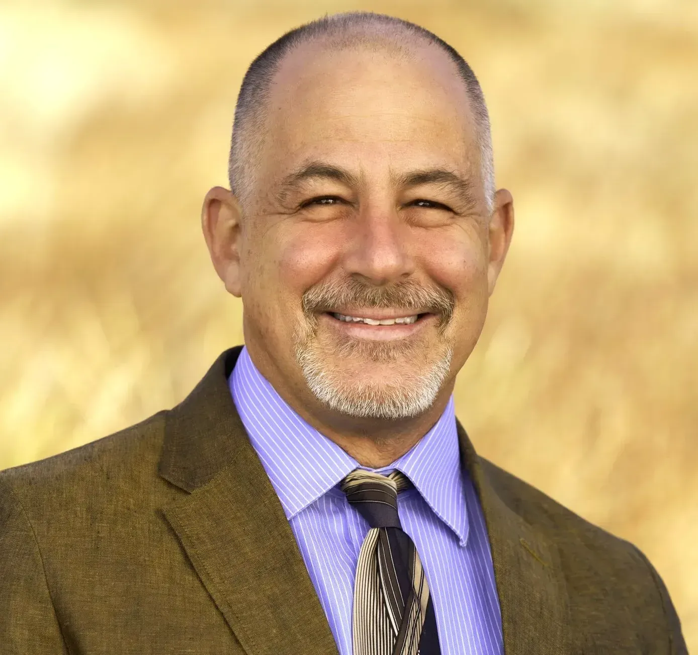 Smiling man, fair skin, short hair, in brown blazer, blue shirt, tie; blurred yellow background.