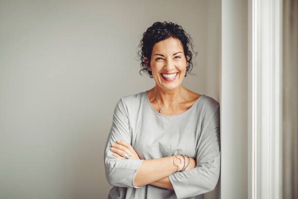 Woman with curly brown hair, smiling, arms crossed, leaning against a white wall.