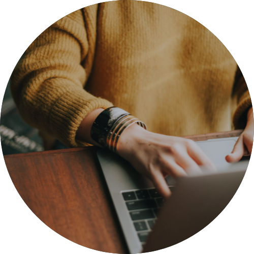 Hands typing on a laptop at a wooden table, wearing a tan sweater and bracelets