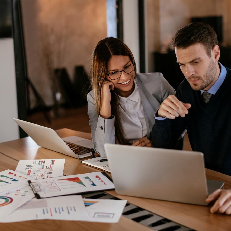 Two coworkers reviewing charts at a laptop in a bright office, one smiling and one pointing.