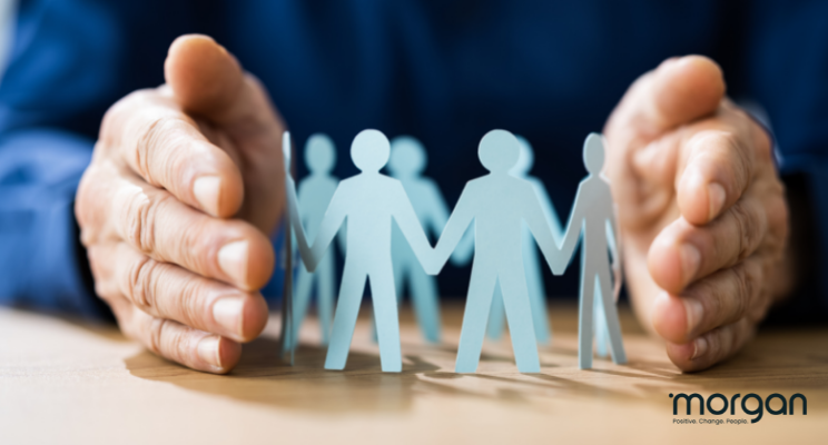 Hands protecting paper family figures on a wooden table, symbolizing care or insurance