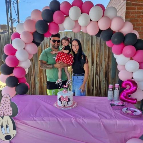 Birthday table with pink-and-black balloon arch, Minnie decor, and two people posing with a child.