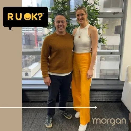 Two people standing indoors by a large window, smiling, with a “R U OK?” speech bubble.