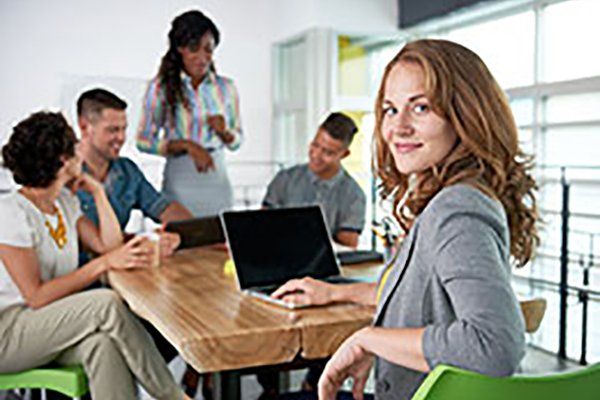 Team meeting in a bright office, with a woman at a laptop in the foreground and colleagues talking nearby