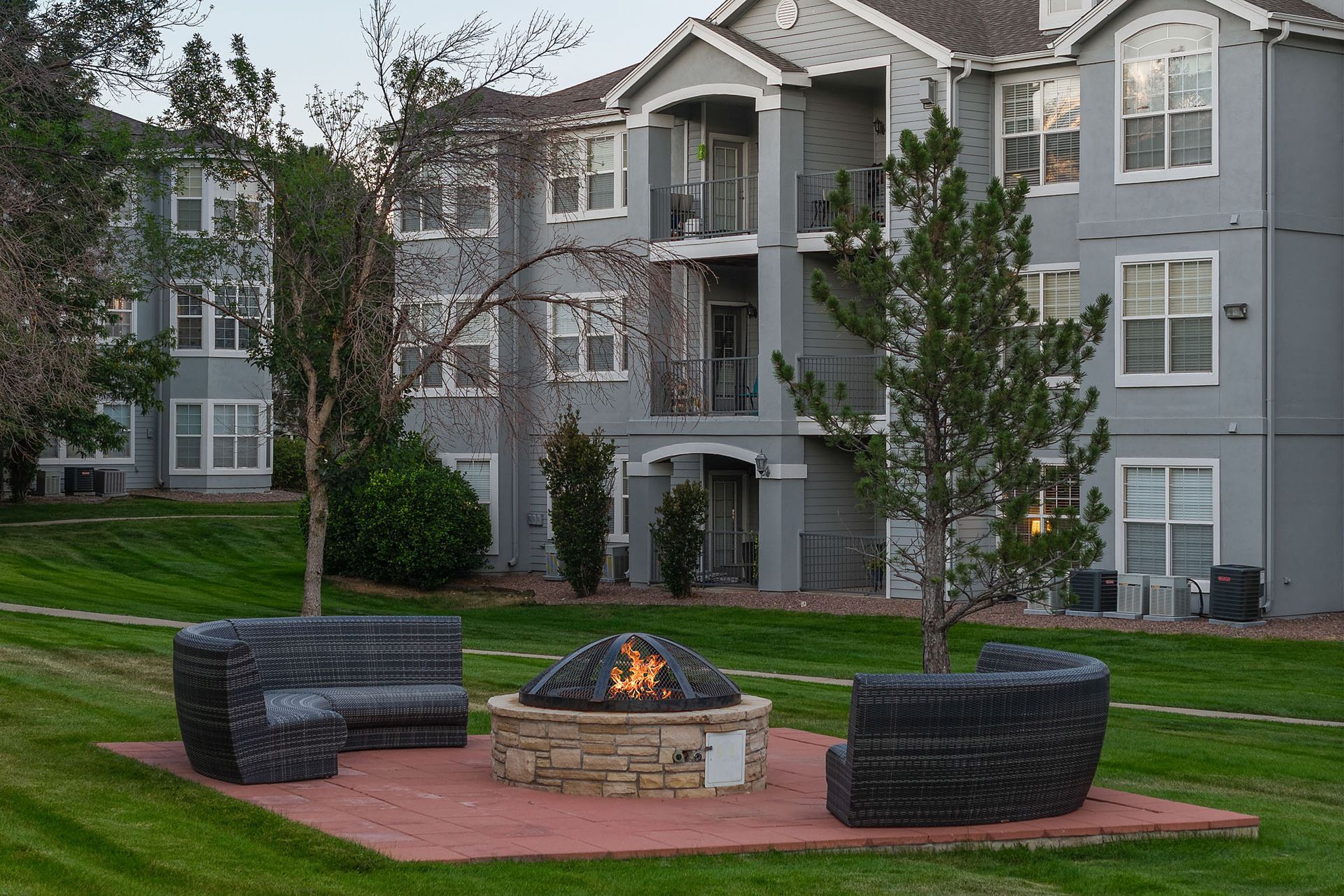 Outdoor fire pit with two dark wicker chairs in front of gray apartment buildings and trees at Marquis at Town Centre in Broomfield, CO