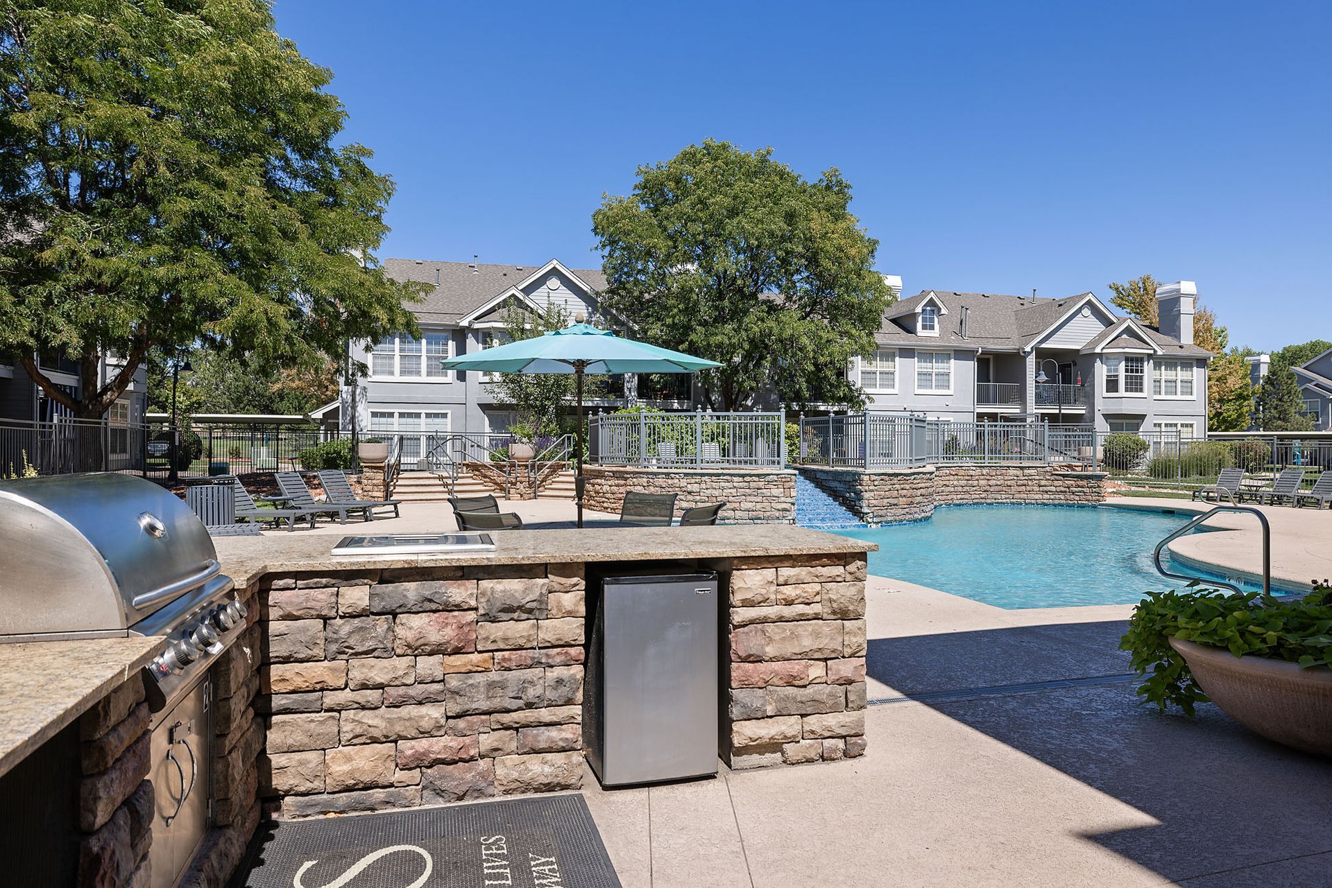 Outdoor pool area with stone patio, grill, picnic tables, trees, and resort-style condos in the background at Marquis at Town Centre in Broomfield, CO