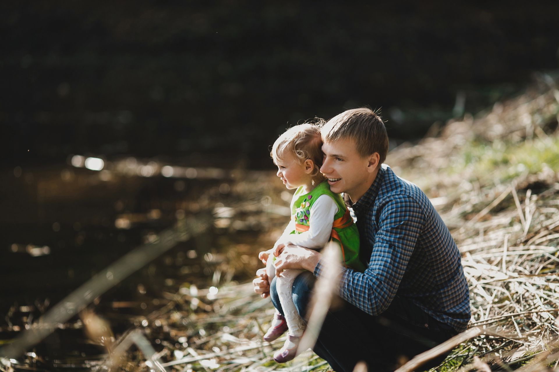 Man sitting by a creek holding a smiling child in a green shirt, with sunlit water and grass behind them