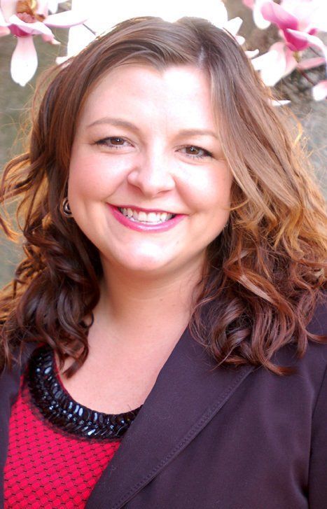 Smiling woman with curly hair in a dark blazer and pink top, standing outdoors with flowers in the background