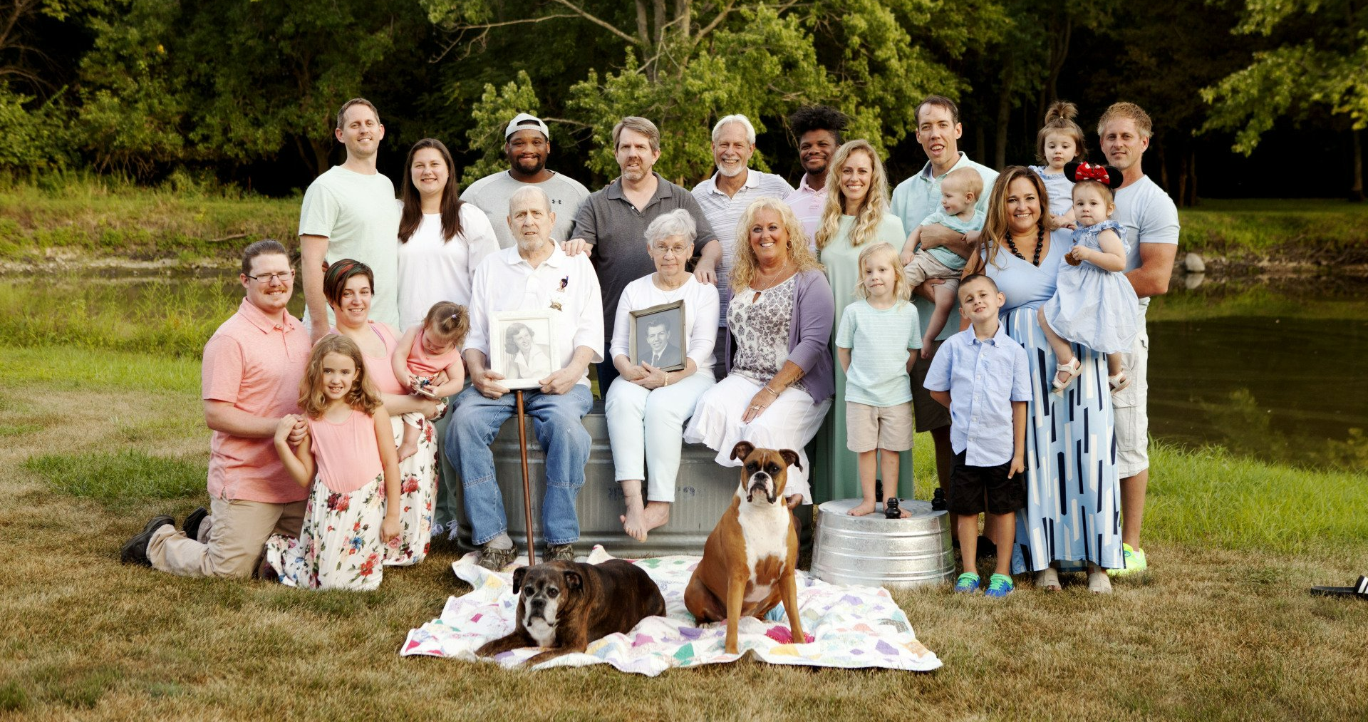 Large family portrait posing outdoors near a pond. Two dogs sit in front, with generations smiling.