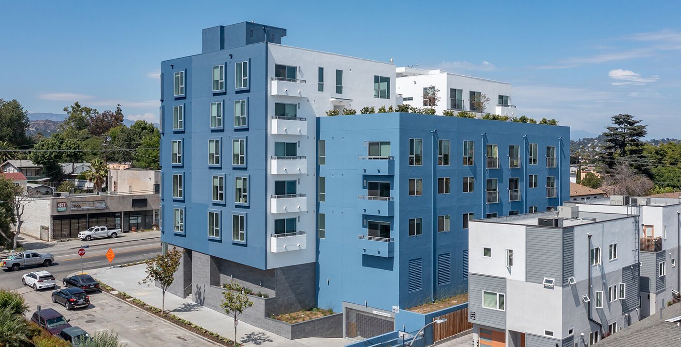 Blue and white apartment building with multiple floors on a sunny day. Cars parked in front on a street.