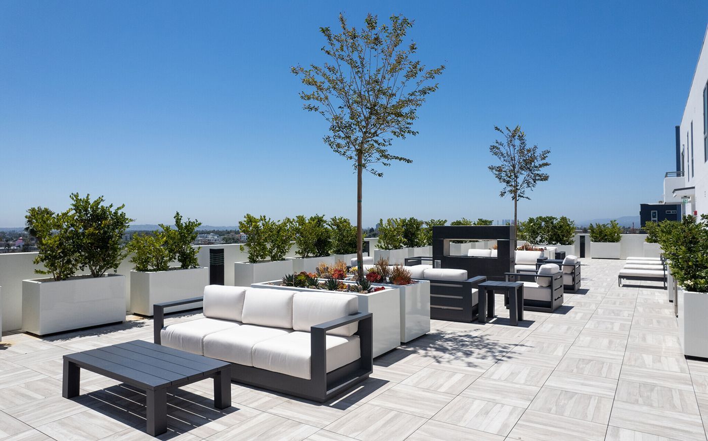 Rooftop patio with white sofas, planters, and a dark table on a sunny day with blue sky.