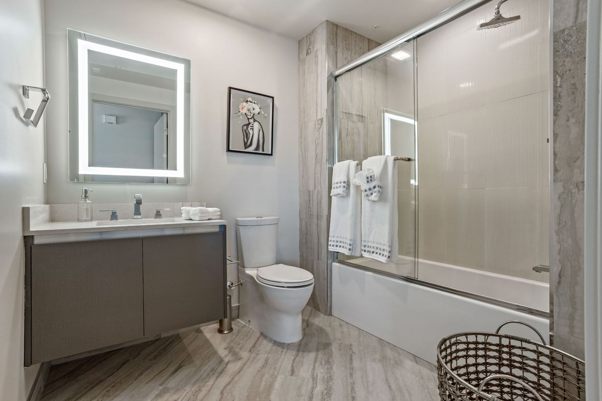 Modern bathroom with gray vanity, backlit mirror, toilet, shower, and a decorative basket.