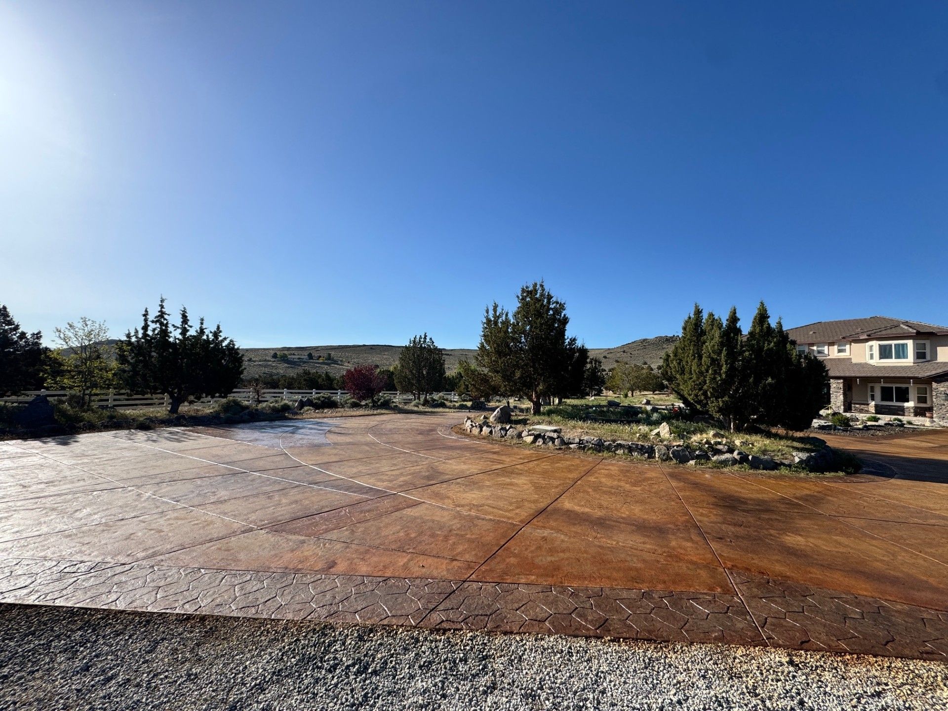 Wide concrete driveway with scattered rocks, trees, and houses under a clear blue sky.