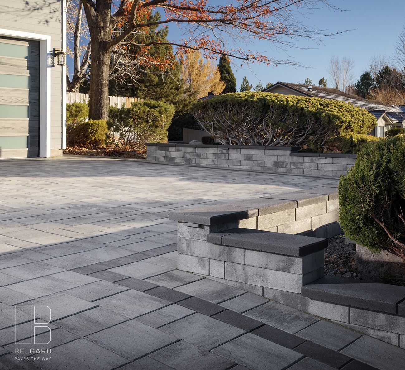 Paved driveway with stone retaining walls and autumn shrubs beside a house under clear daylight