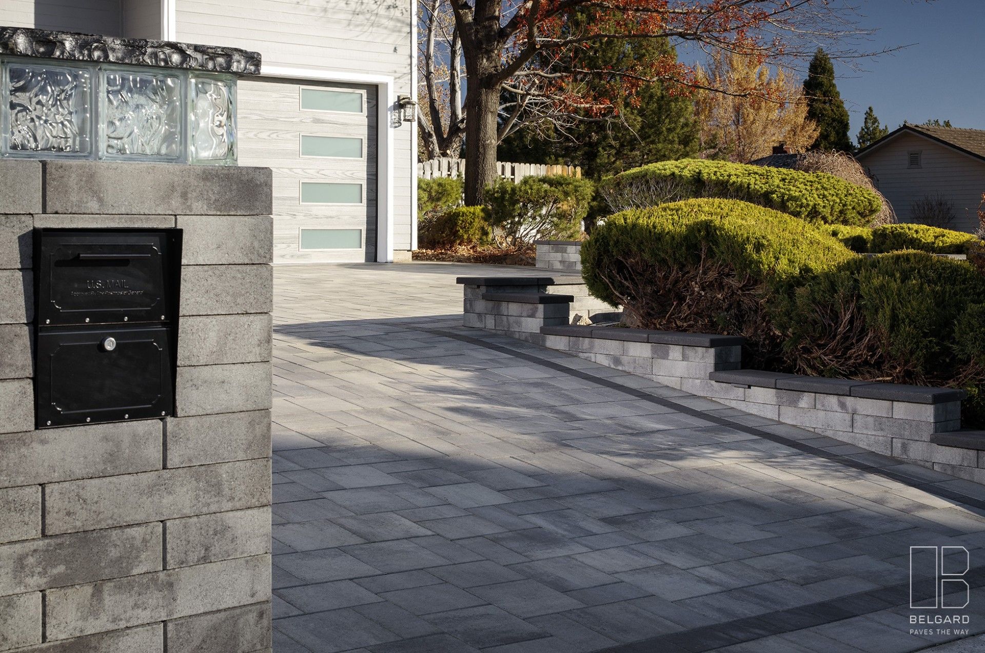 Modern house driveway with gray pavers, stone mailbox pillar, garage door, and landscaped shrubs