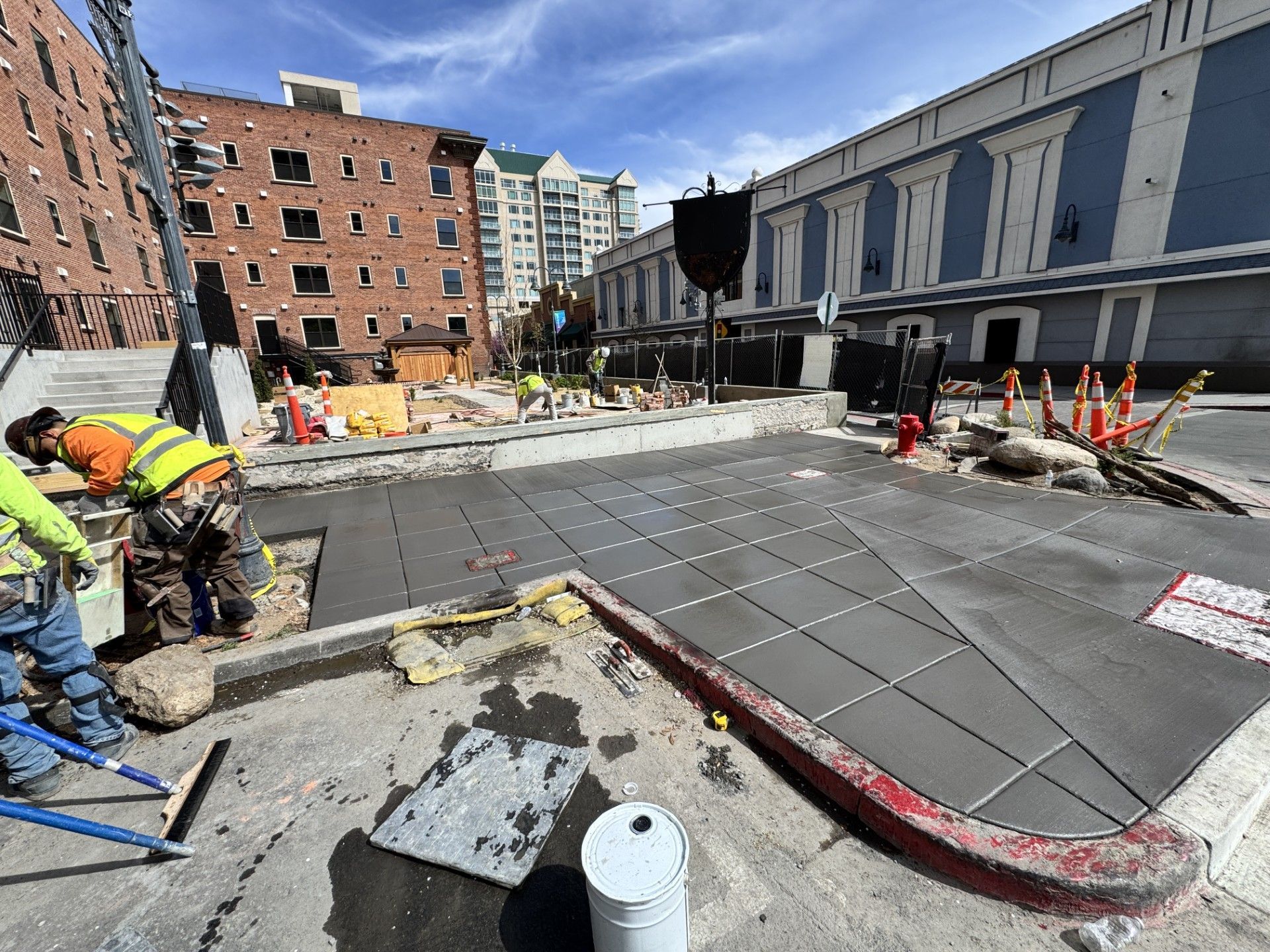 Construction workers paving a sidewalk beside a brick building and street barrier cones