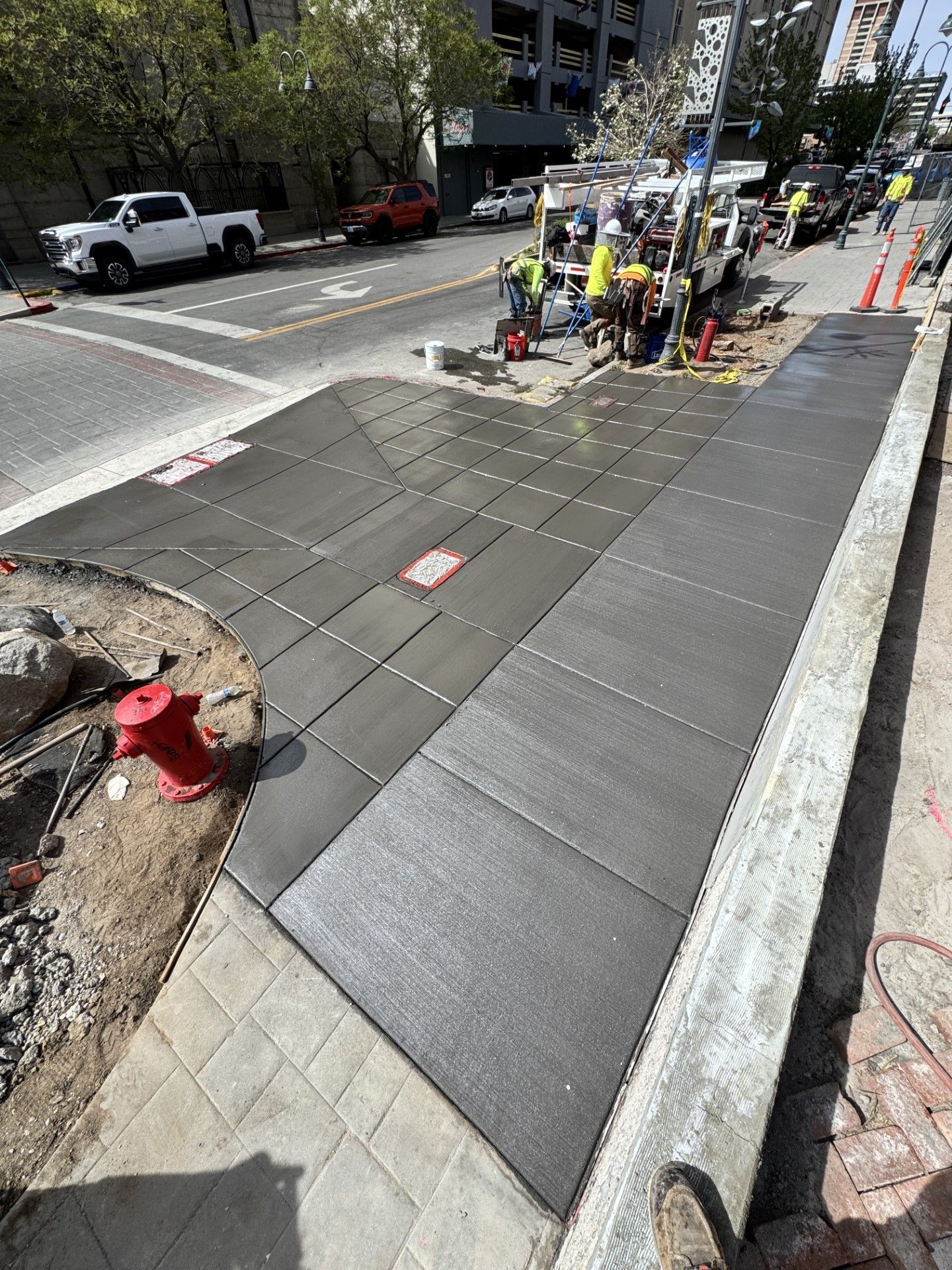 Construction crew installing sidewalk panels along a city street with traffic cones and parked vehicles nearby