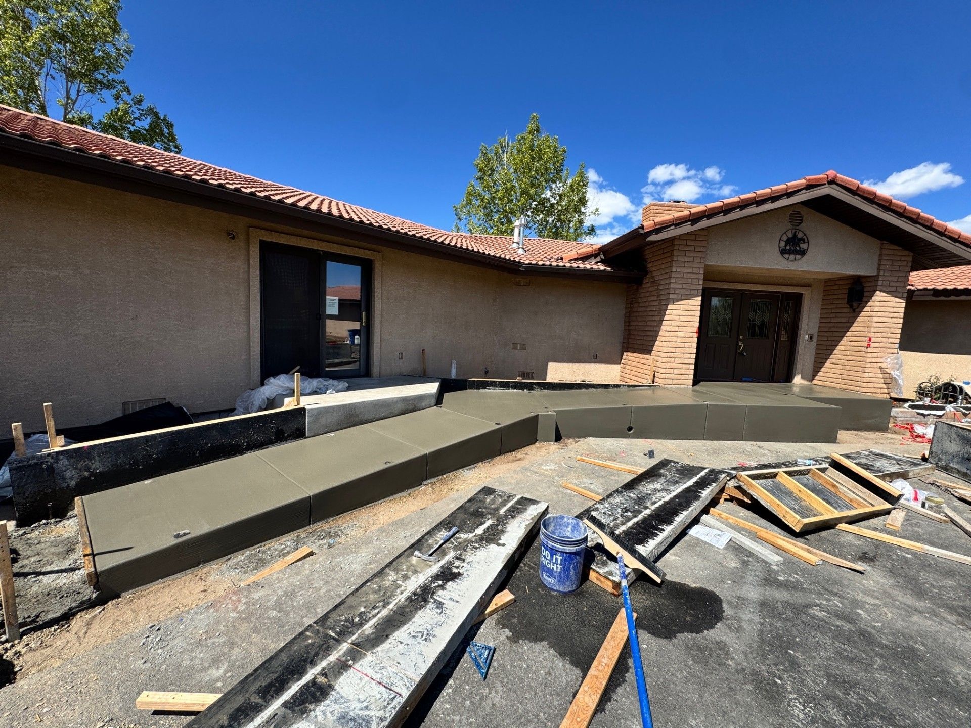 Courtyard under construction with concrete forms, tools, and a stucco house under a blue sky