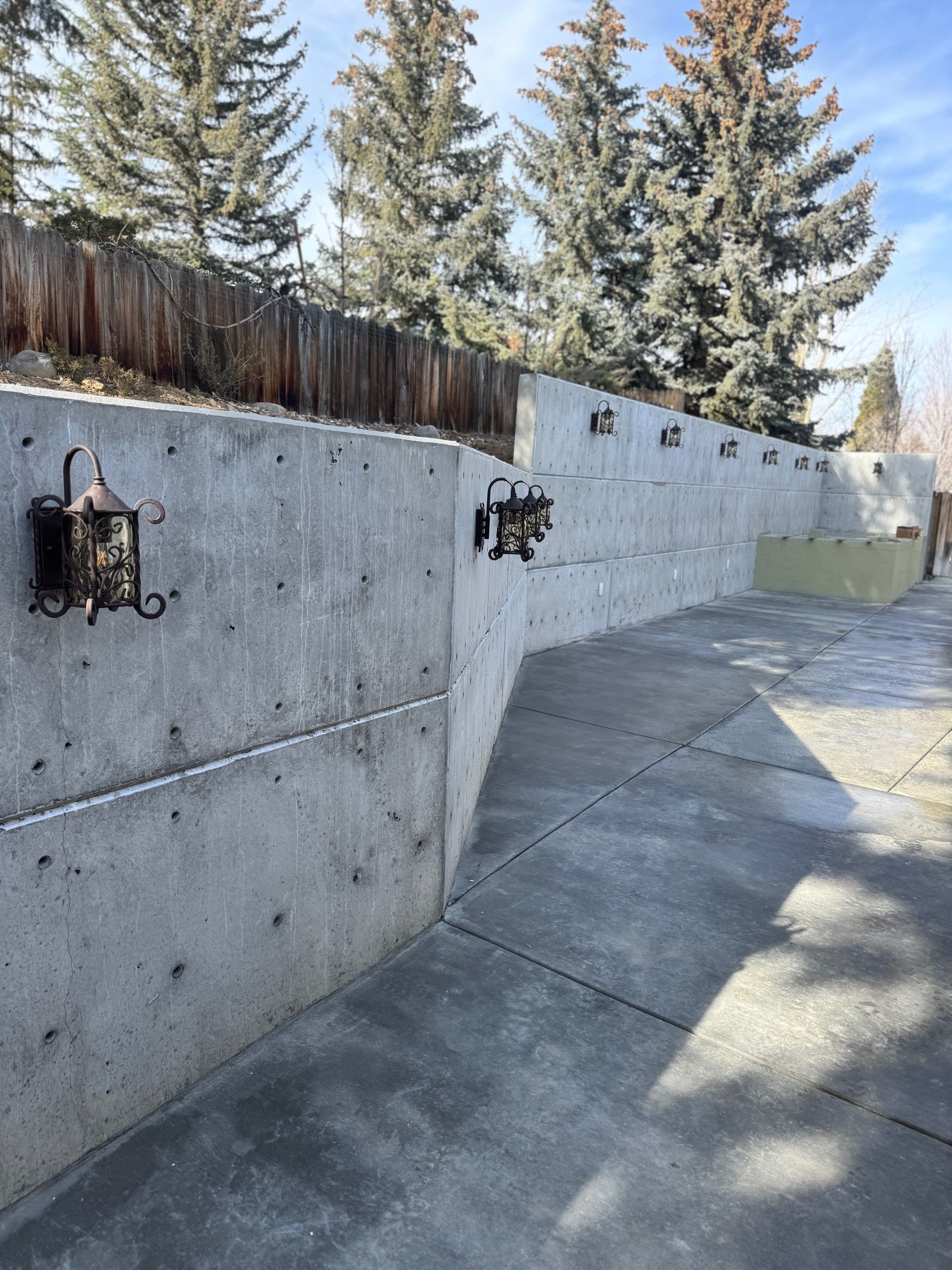 Concrete walkway beside a gray retaining wall with small black lanterns and tall trees above