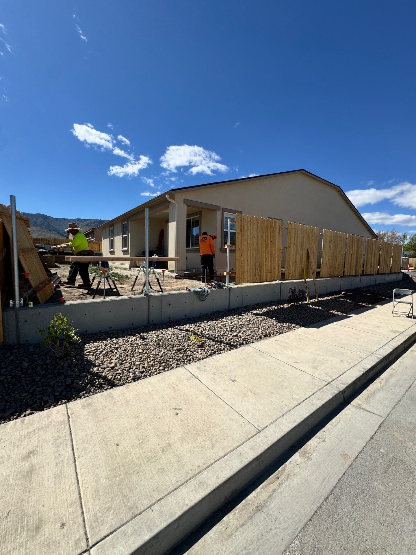 Single-story house under construction with wooden fencing, gravel yard, and a clear blue sky.