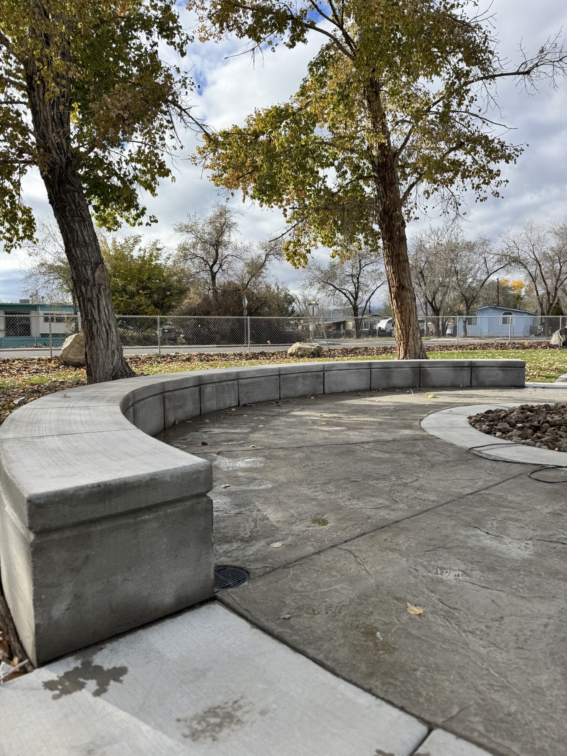 Curved concrete bench in a park with trees and a paved walkway under cloudy skies