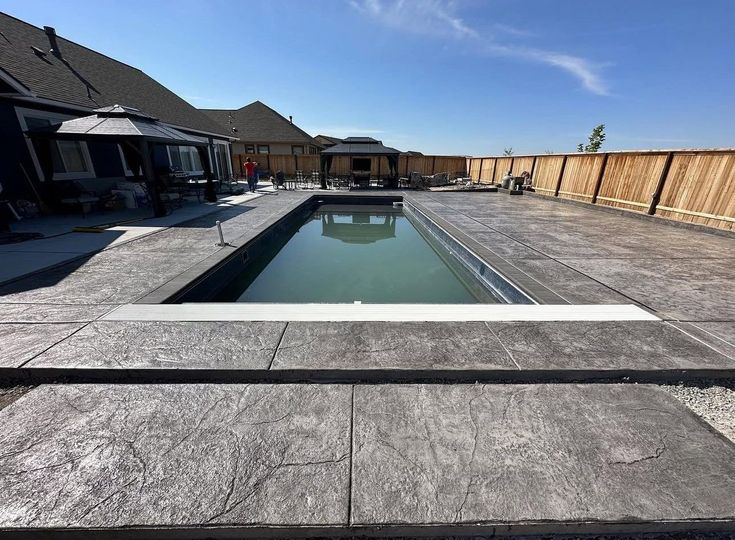 Backyard with rectangular swimming pool, stone patio, and houses under a clear blue sky
