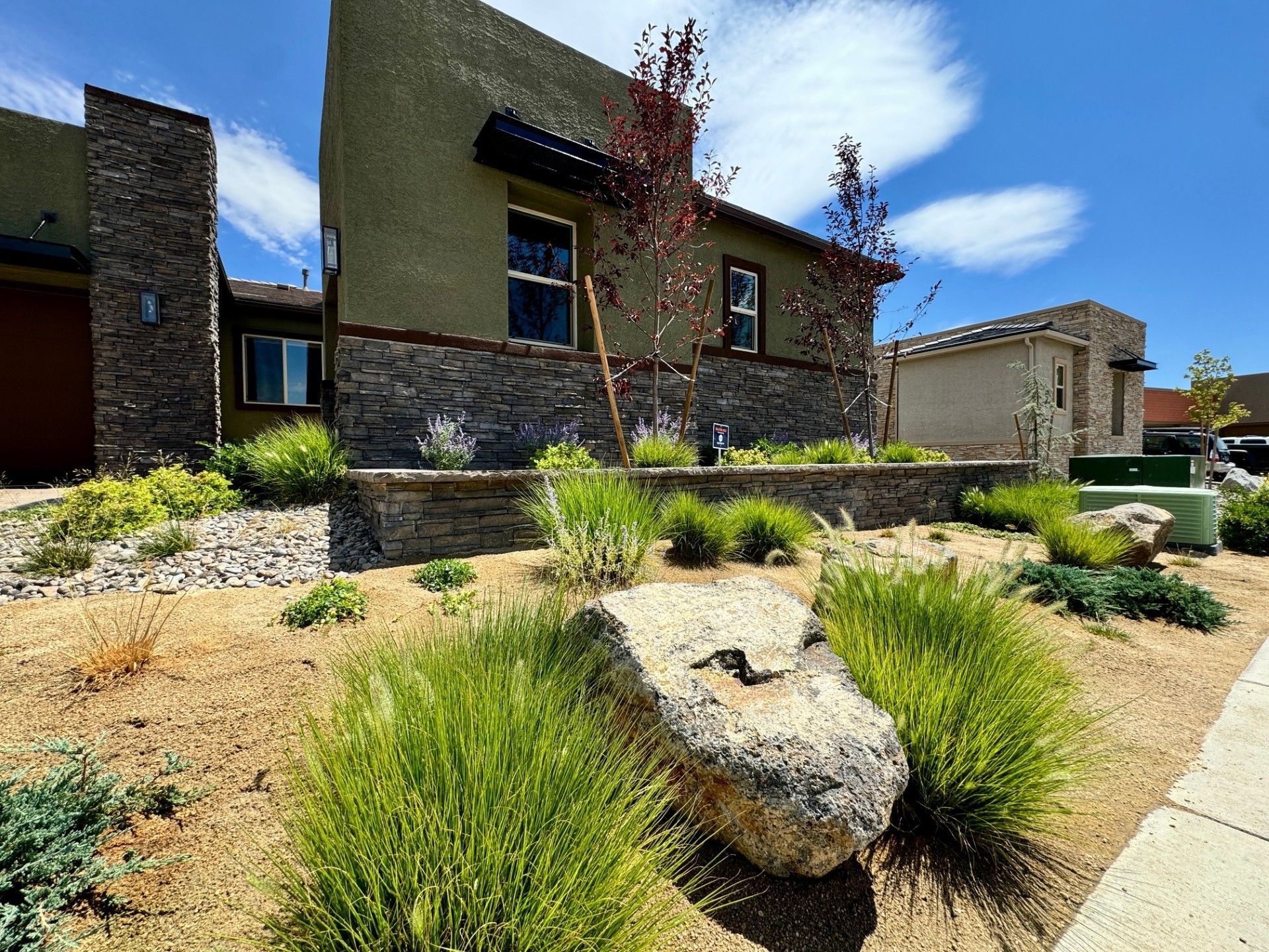 Modern house with landscaped desert garden, large boulder, shrubs, and sidewalk under a blue sky