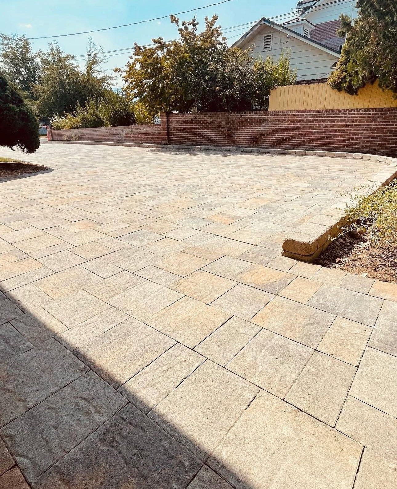 Paved driveway beside a brick wall and house, with trees and bright sunlight.