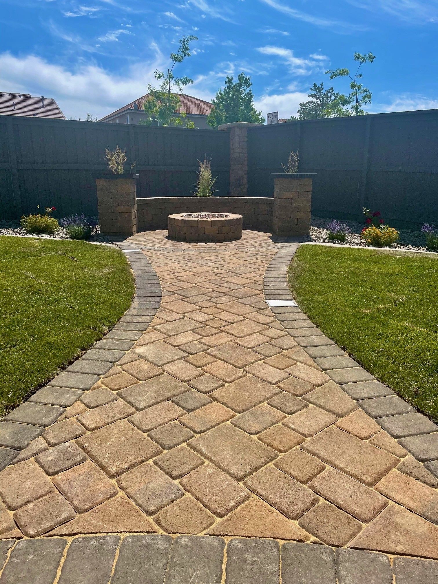 Curved stone walkway leading to a circular fire pit in a landscaped backyard under a blue sky