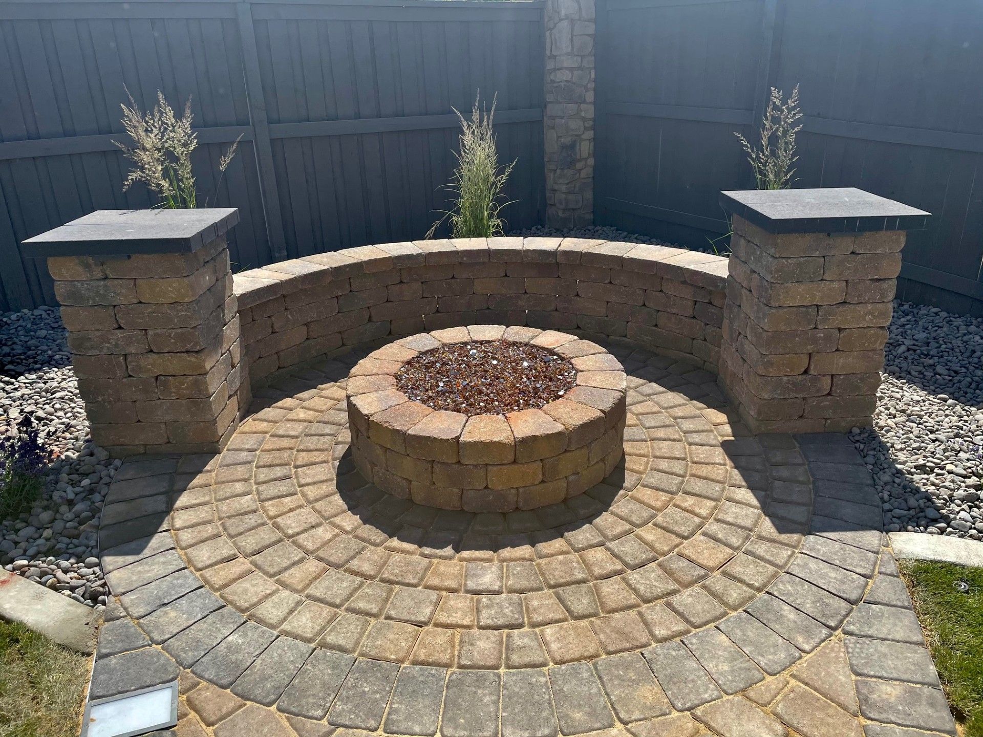 Circular brick fire pit with curved stone bench and two square brick pillars in a backyard patio