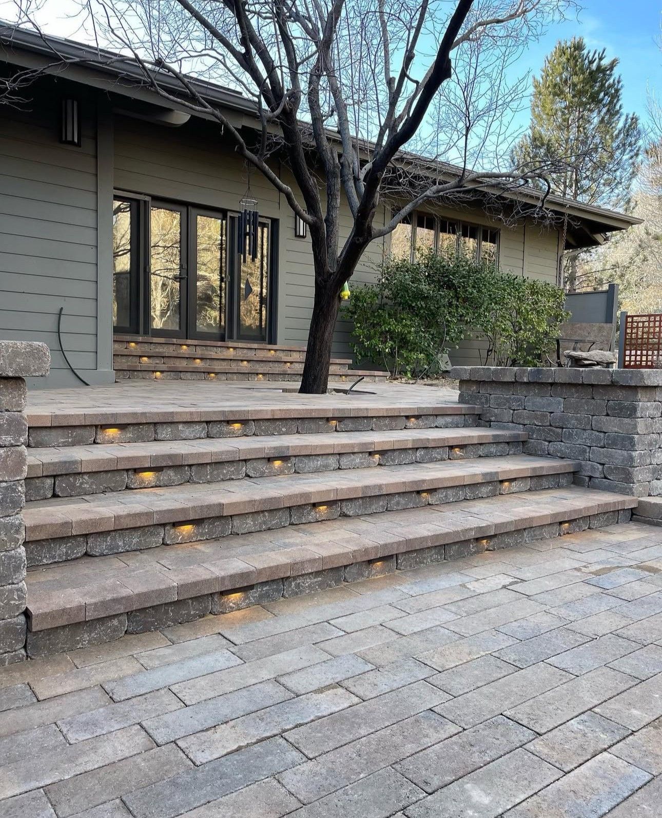 Stone steps lead to a house entrance with large windows and a tree in front of the patio.