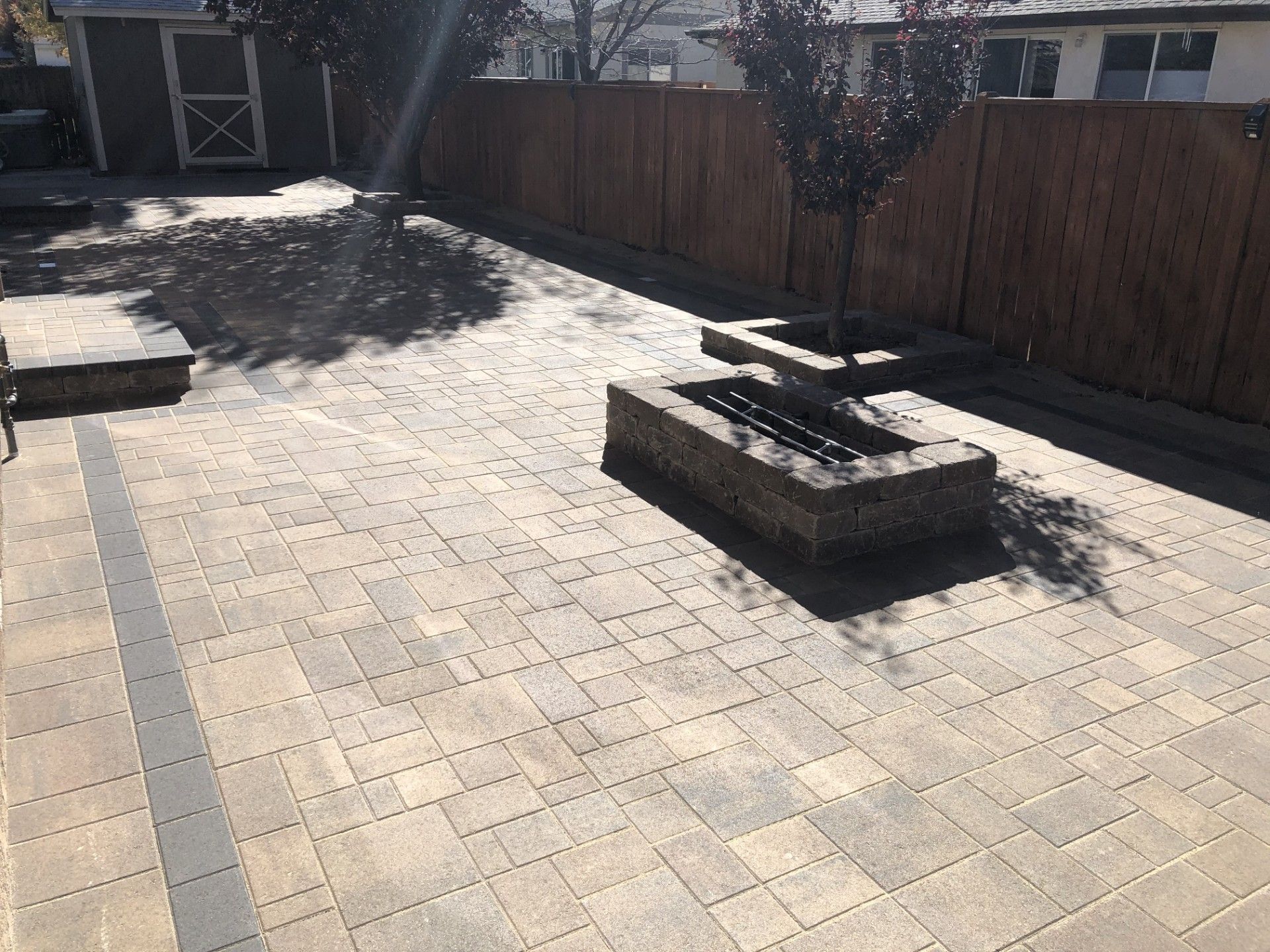 Sunlit backyard patio with pavers, a brick fire pit, wooden fence, and a small bench