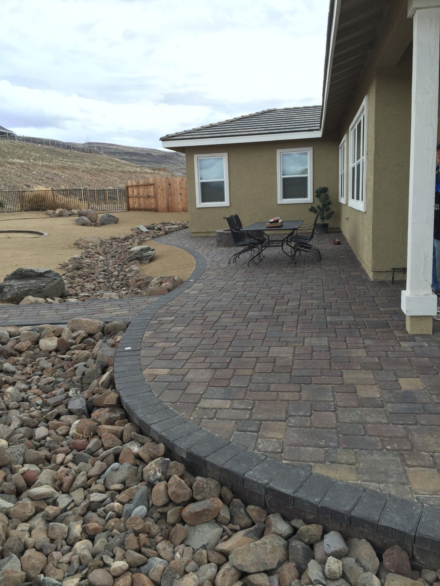 Curved brick patio with a small table and chairs beside a beige house and rock landscaping