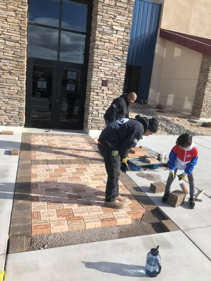 Two workers install brick pavers on a front walkway outside a stone house.