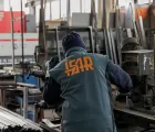 Un operaio con una giacca color verde acqua aziona dei macchinari in un'officina metallurgica, circondato da attrezzi e attrezzature.