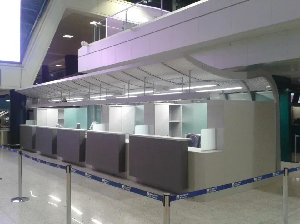 Banchi del check-in aeroportuali vuoti, con divisori grigi e paletti blu sotto una luminosa passerella sopraelevata.