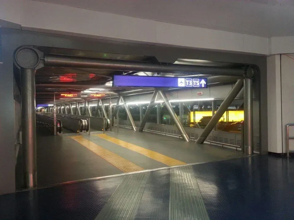 Ingresso del terminal aeroportuale con porte a vetri, travi metalliche e un'insegna luminosa soprastante.