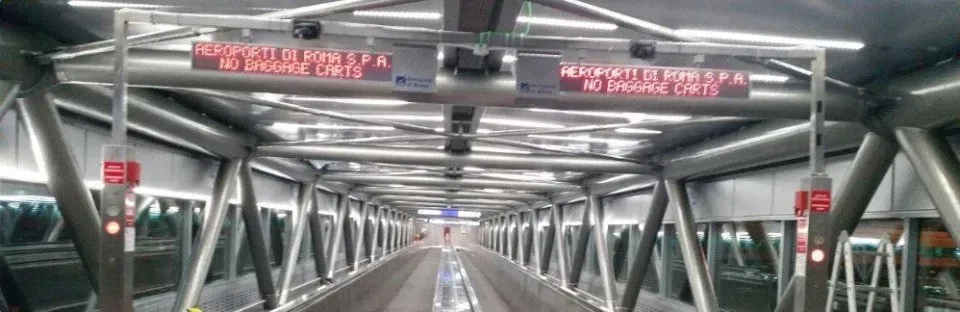 Interno di un ponte pedonale metallico con travi ripetute e segnali rossi in alto