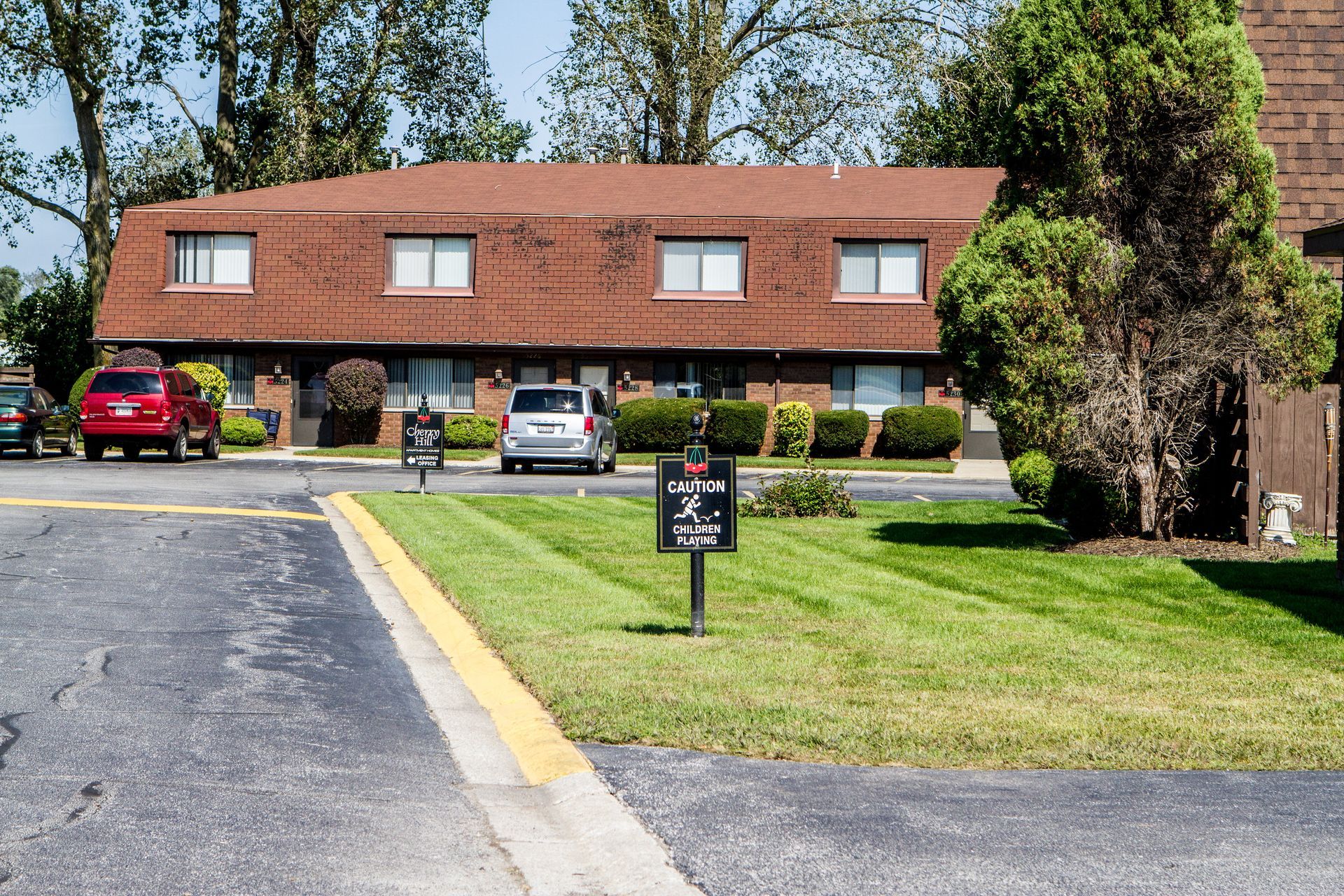 Brown roofed apartment building with a grassy lawn in front, cars parked nearby.