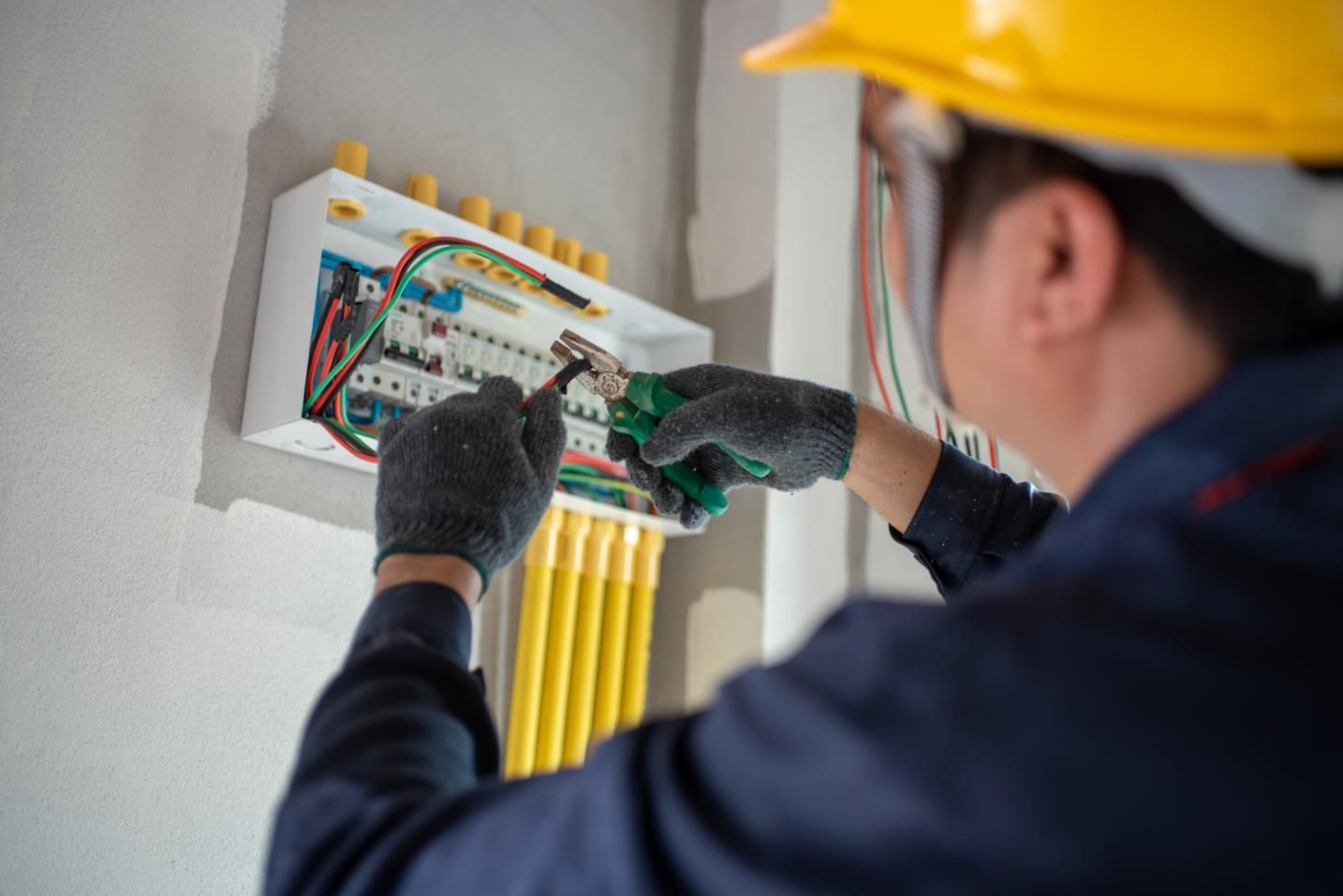 Electrician working on electrical panel, wearing gloves and hard hat.
