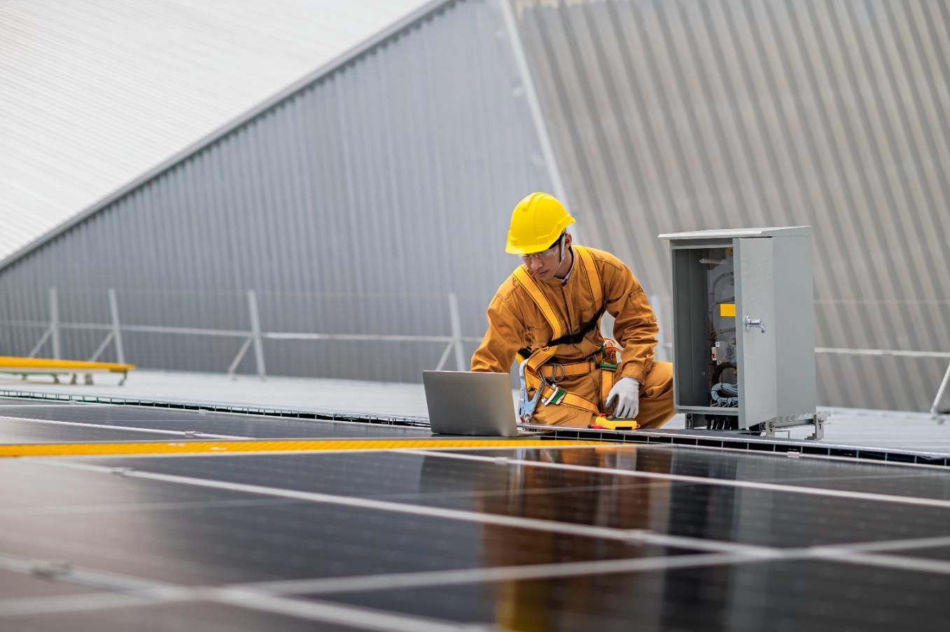 Worker in yellow hard hat and jumpsuit on rooftop inspecting solar panels with a laptop.