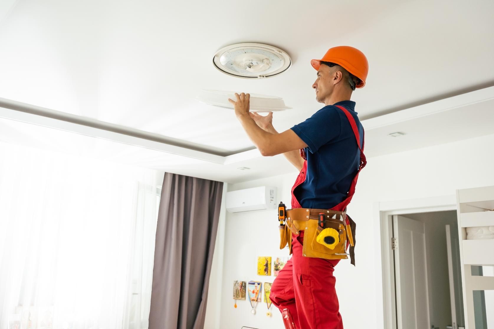 A worker in an orange hard hat installing ceiling light fixture in a room; standing on a ladder.