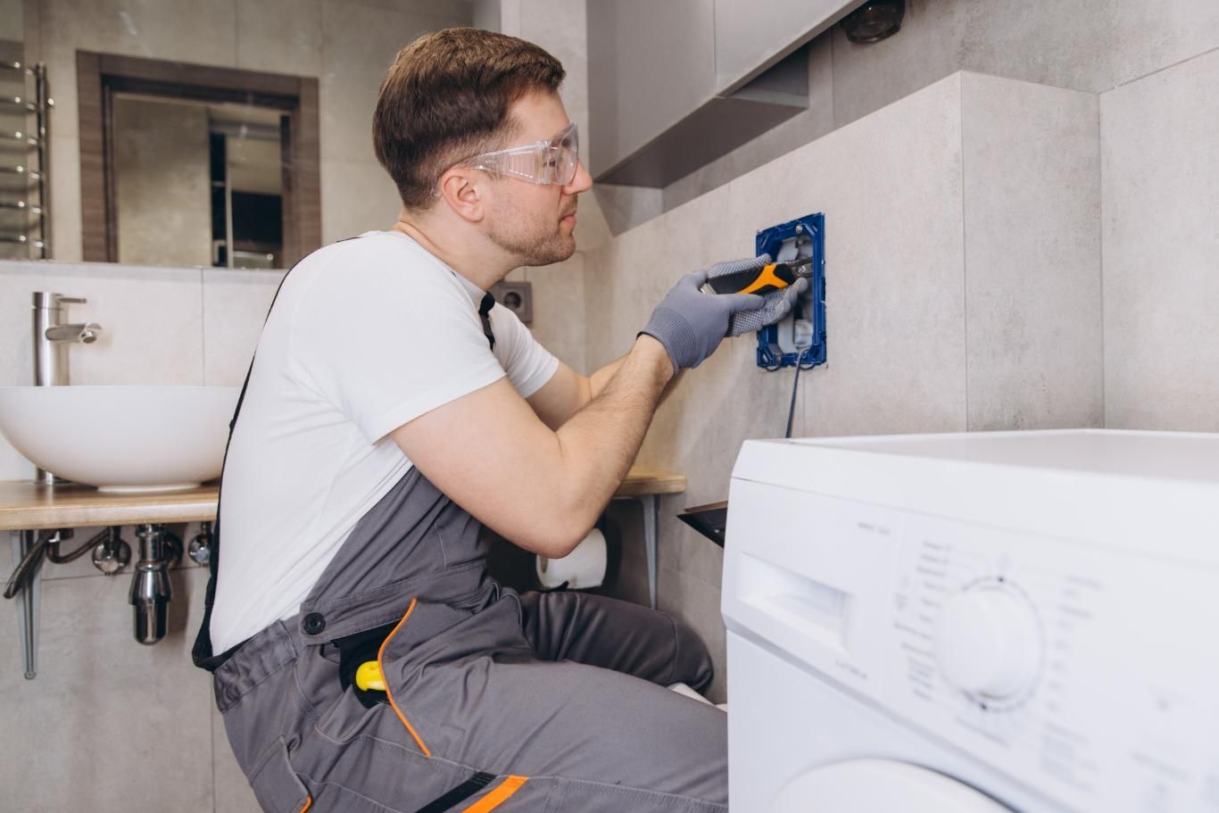Man in overalls and safety glasses wiring outlet in a bathroom, near a washing machine and sink.