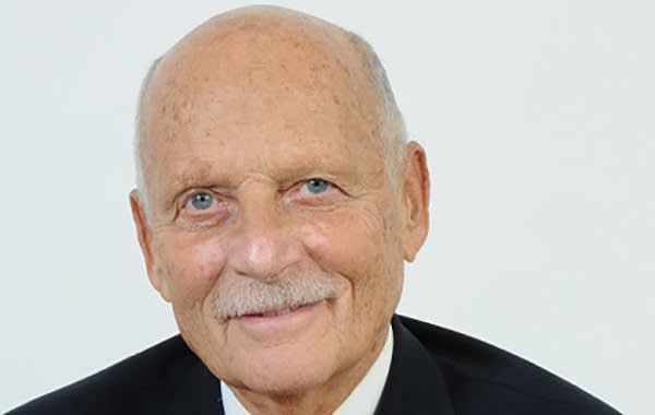 Portrait of a bald man with a gray mustache in a black suit, smiling against a light background