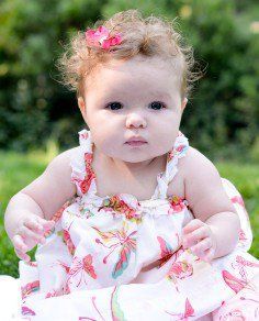 Baby in a white floral dress with a pink flower headband, sitting outdoors with a neutral expression