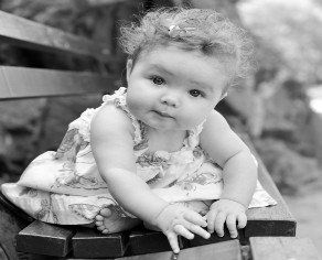A baby in a patterned dress crawling on a wooden bench, looking toward the camera.