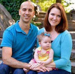 Family of three sitting outdoors on a bench, smiling, with a baby in a pink outfit in front.